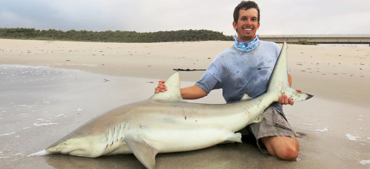 Josh Jorgensen kneeling on shore with a shark that he caught